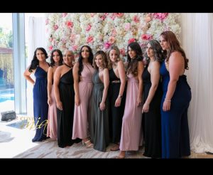 9 women wearing full length gowns pose in front of a white and pink flower wall.